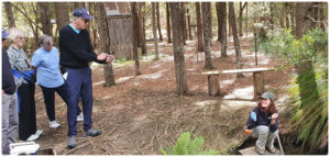 Young Staff member crouched at edge of enclosure while group of older visitors listen to her presentation. One gentleman is standing forward with his phone taking a picture of the staff member.